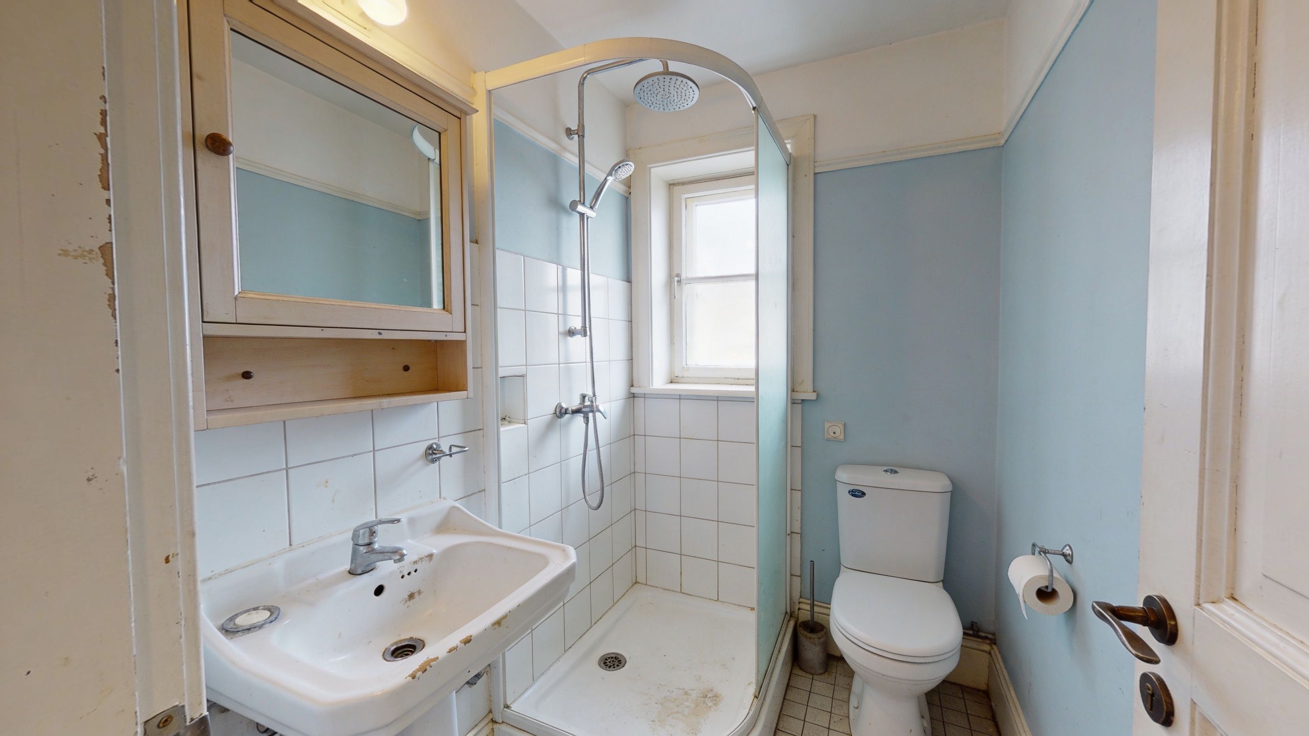 Et ældre badeværelse med behov for renovering, der indeholder en håndvask, en brusekabine og et toilet.